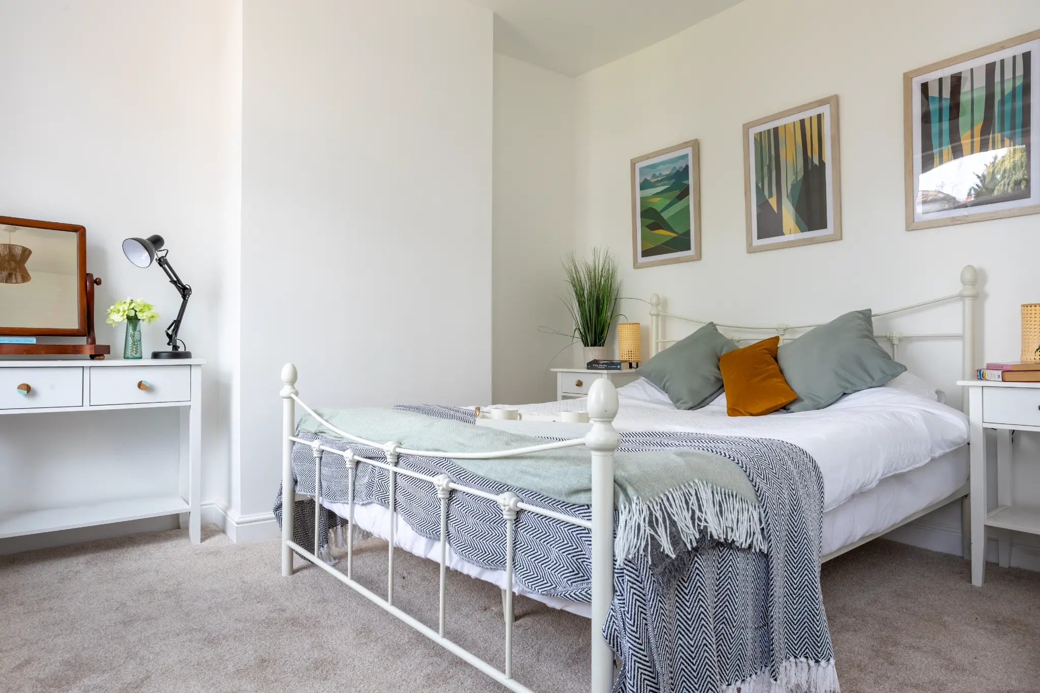 Guest bedroom with white metal bed frame, sage and mustard cushions, and landscape art prints