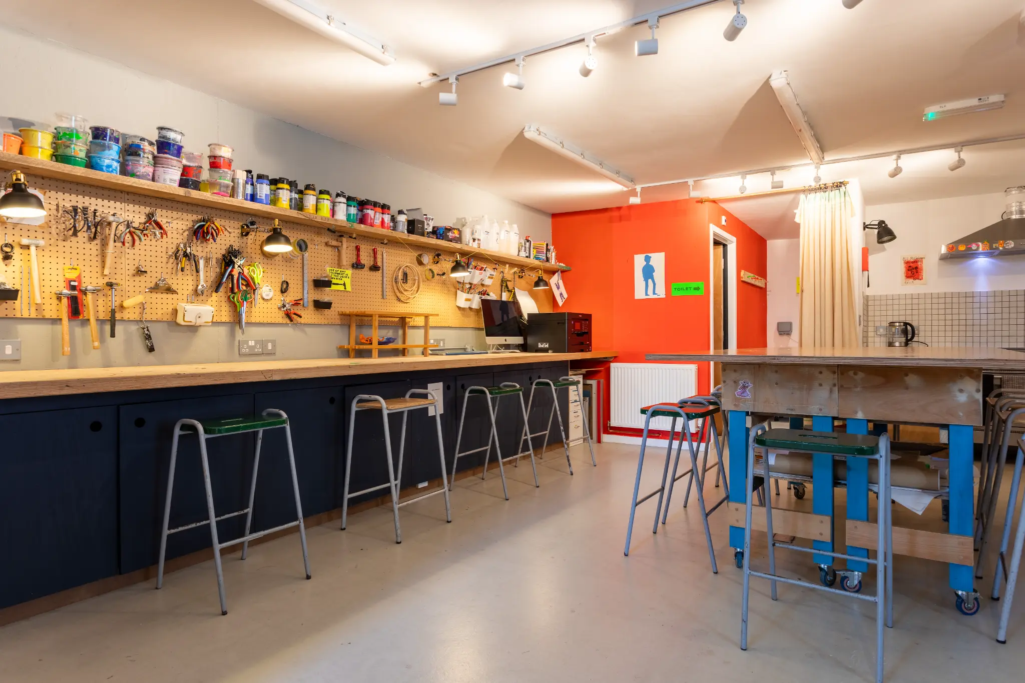 Workshop space with pegboard tools, paint supplies, workbenches, stools, and orange accent wall