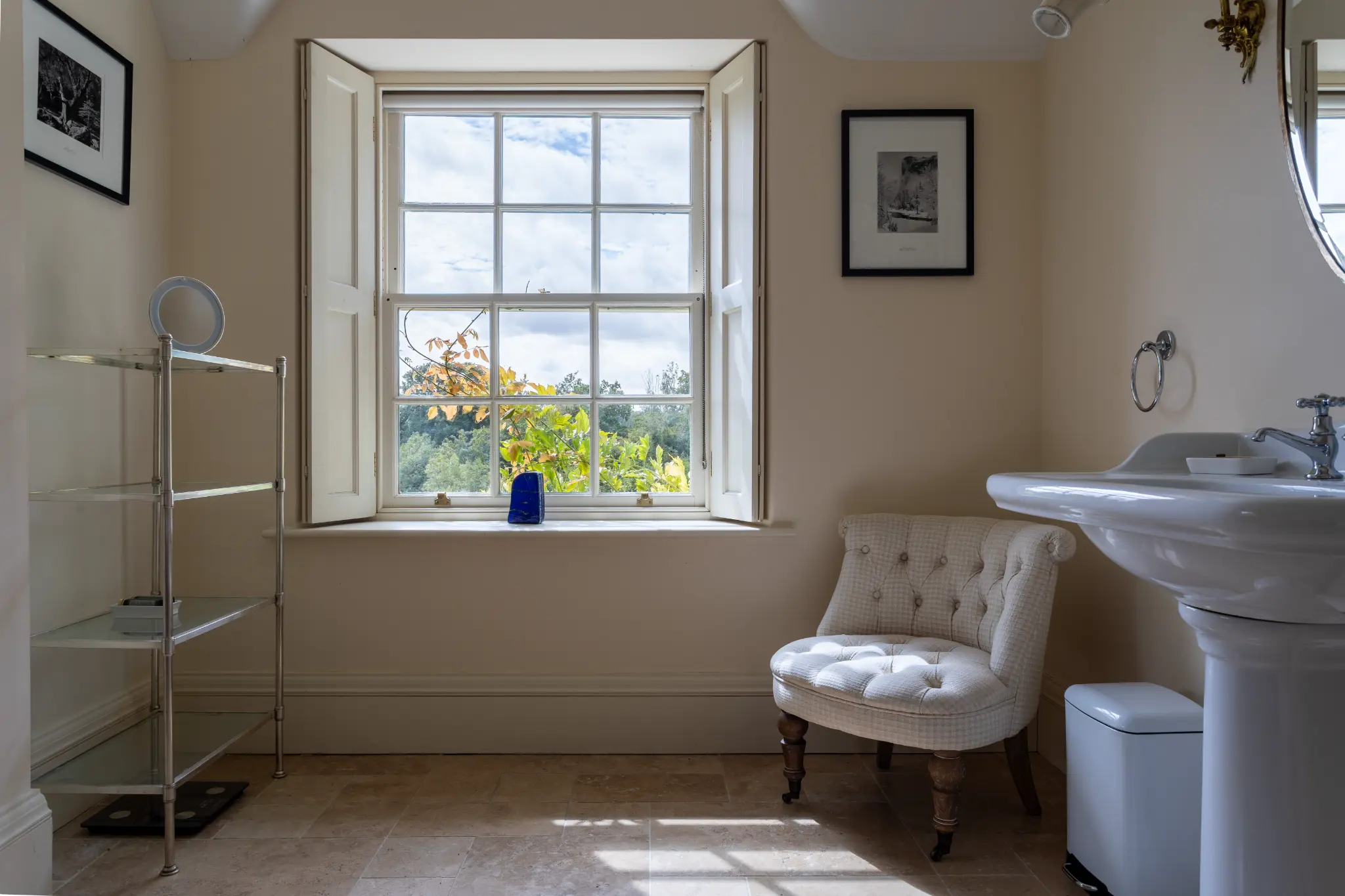 Bright and elegant bathroom featuring traditional wooden window shutters, a classic pedestal sink, a tufted armchair, and scenic countryside views