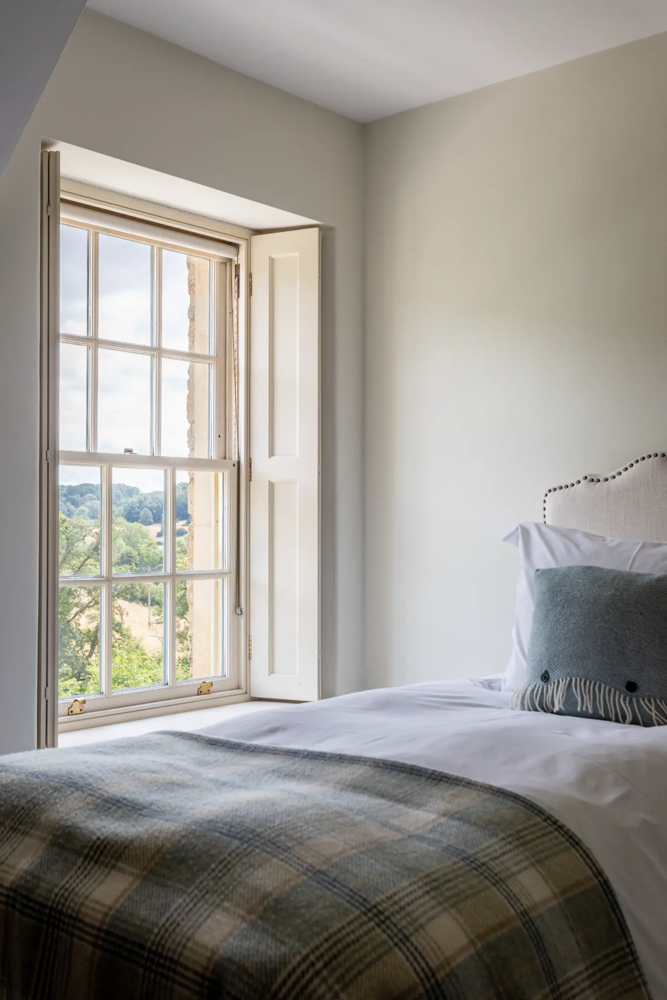 Charming bedroom featuring a tall sash window with interior shutters, neutral upholstered headboard, and tranquil views of the English countryside