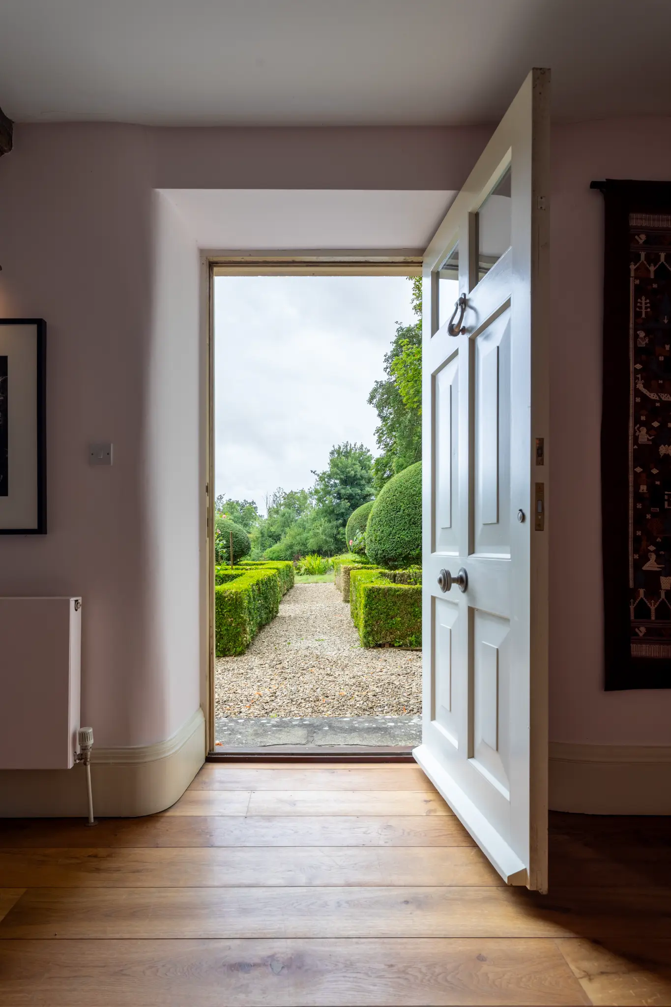 Open white panelled door from a historic manor hallway looking out to a formal gravel garden with sculpted topiary hedges