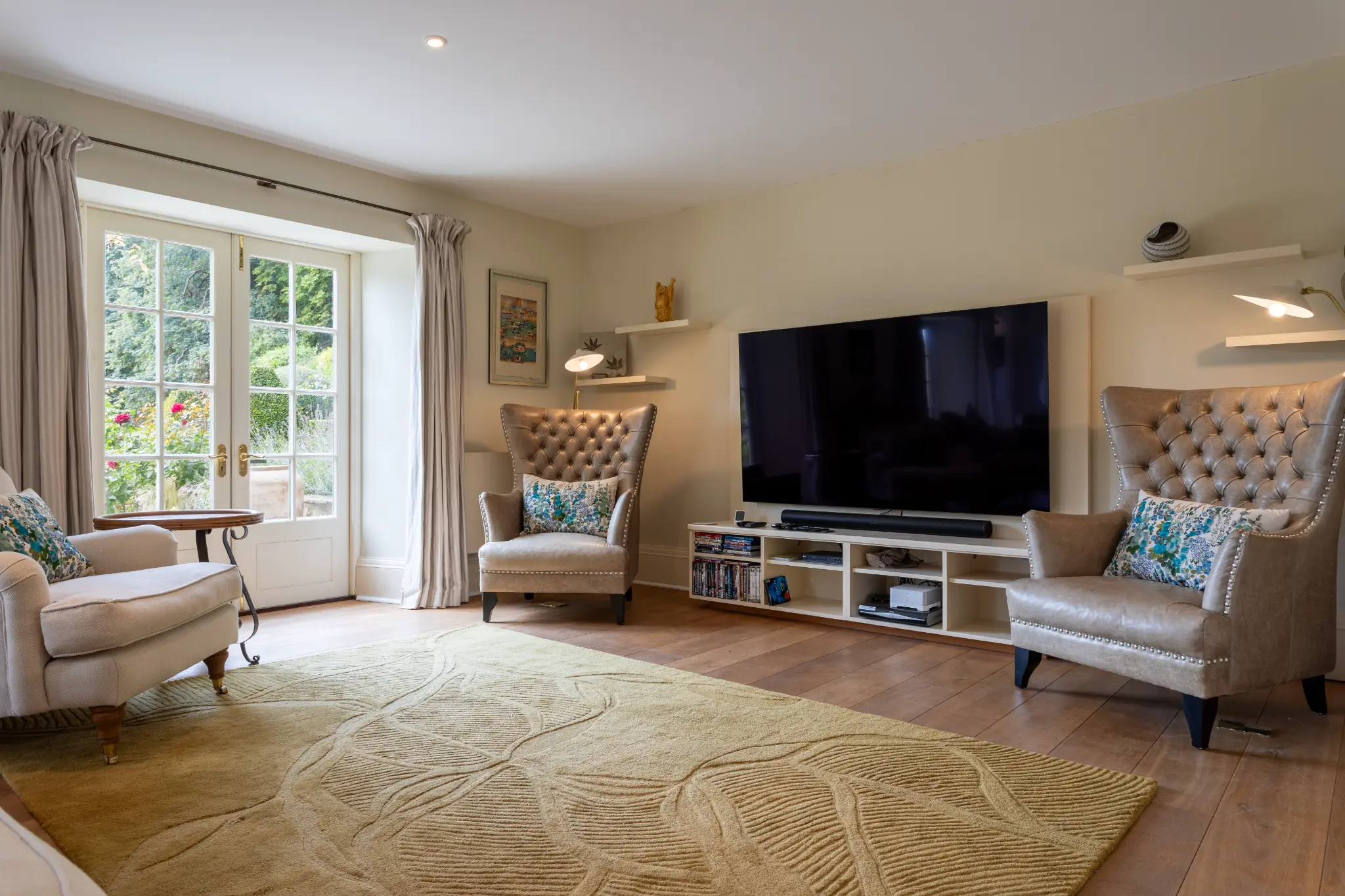 Comfortable living room at a luxury Bath manor featuring plush tufted armchairs, a large television, and garden views through French doors