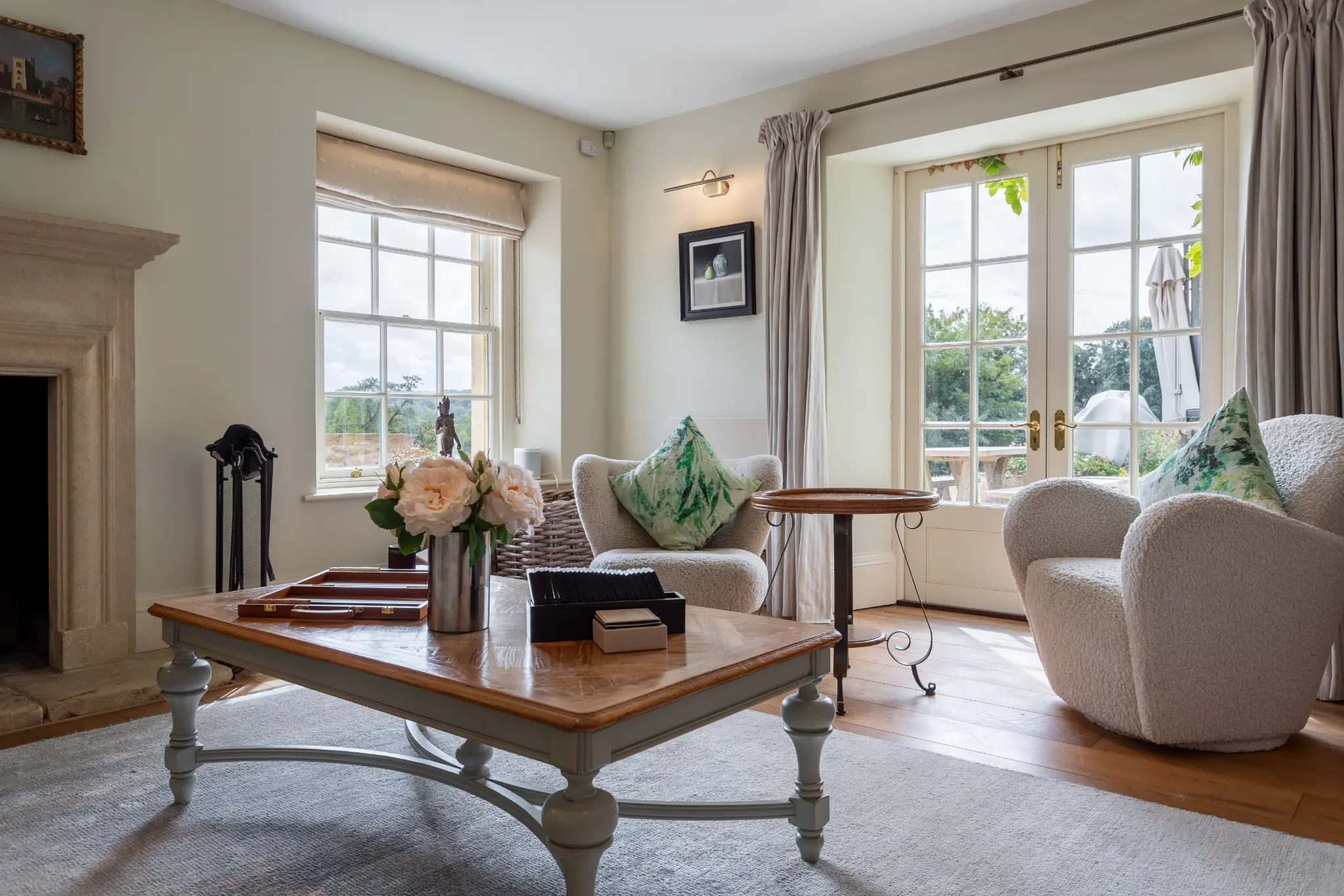 Cozy living room with plush cream armchairs, a stone fireplace, and French doors opening to the grounds of a luxury Bath manor