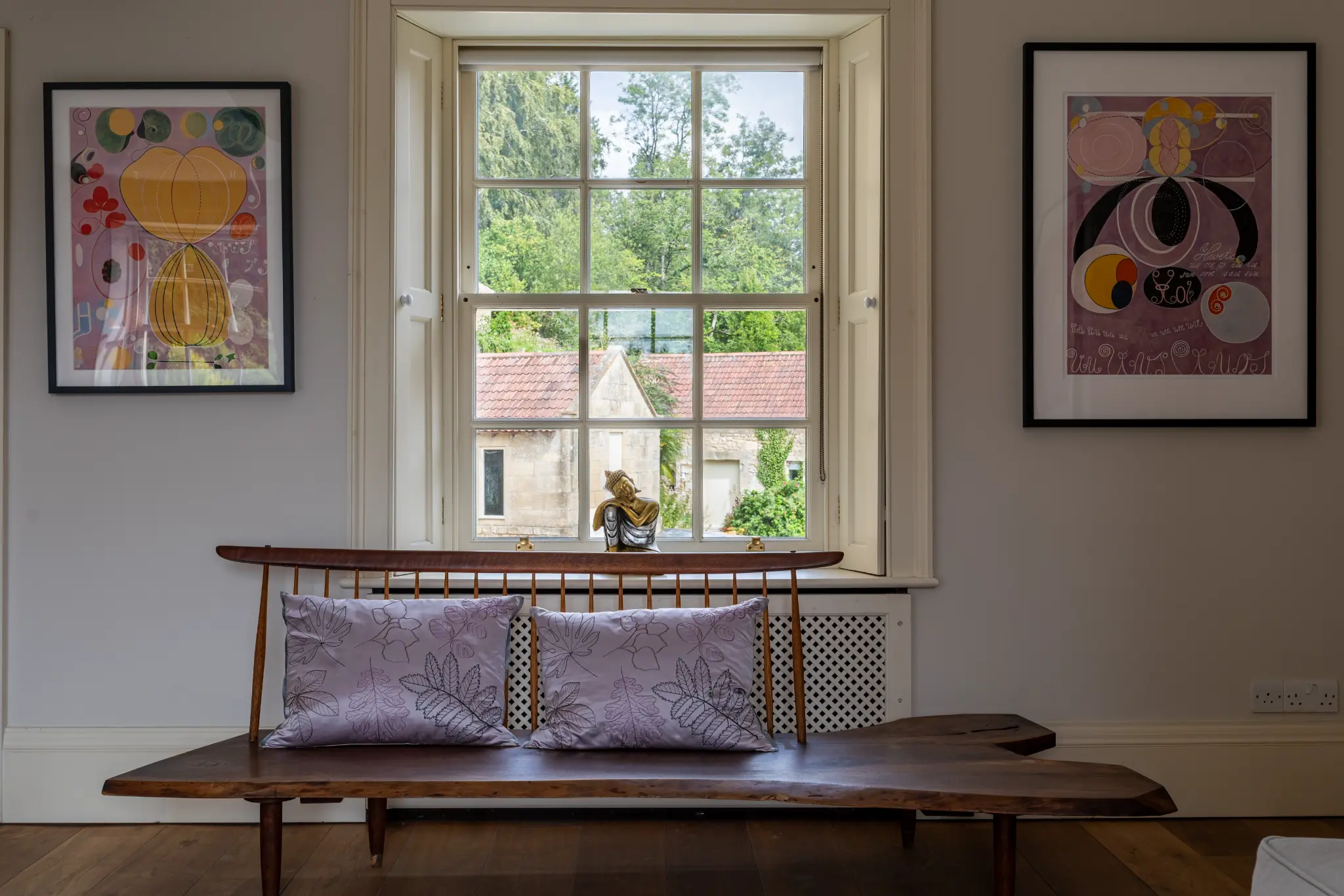 Wooden bench with patterned cushions beneath an open sash window flanked by abstract art in a bedroom at a luxury Bath manor