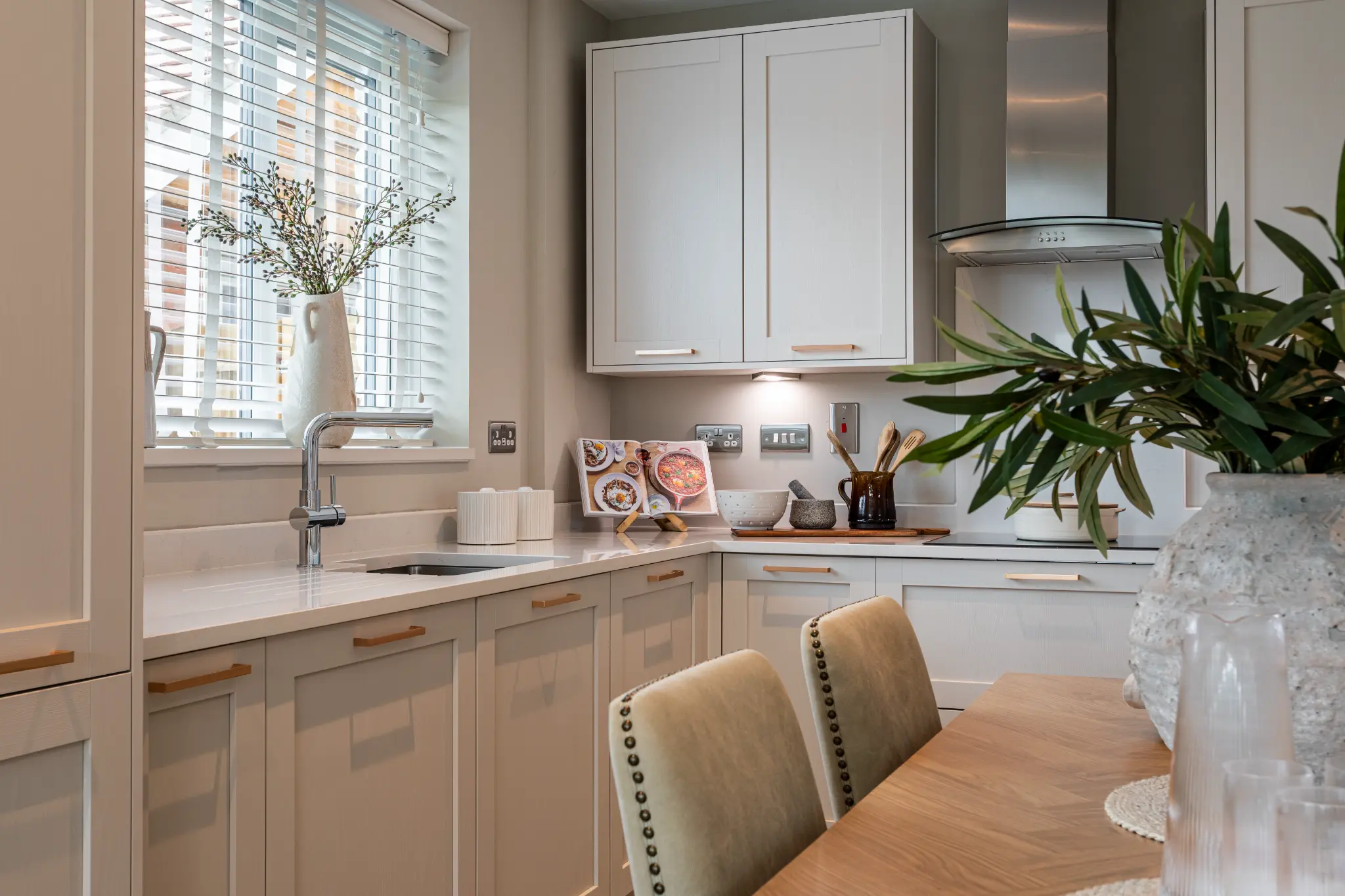 High-end show home kitchen and dining area with pale cabinetry, natural timber and layered greenery