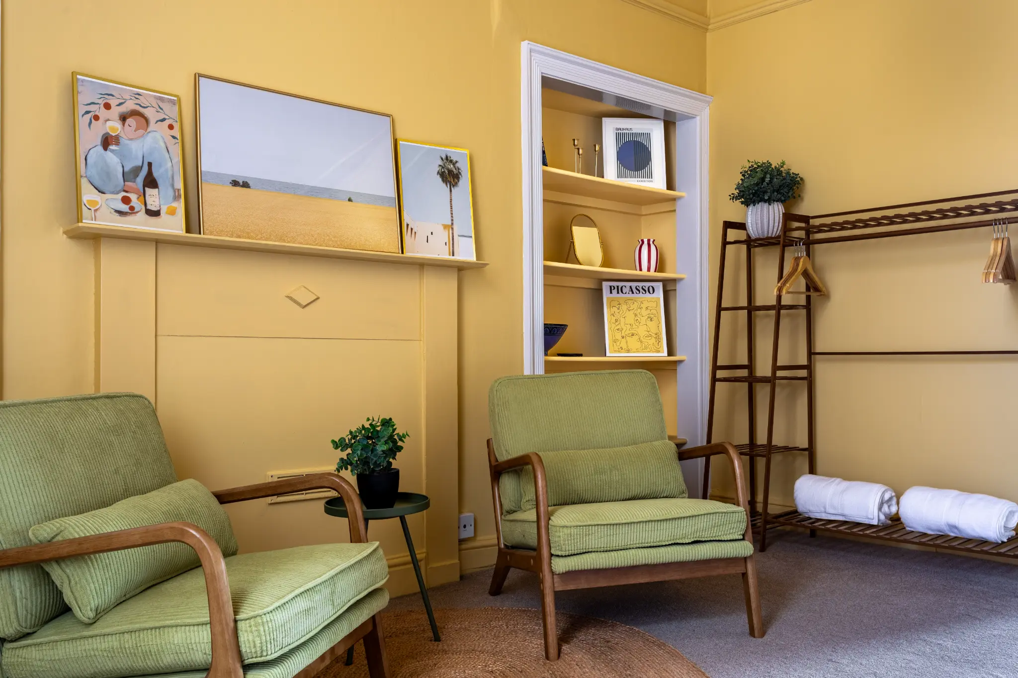 Amber bedroom corner of a Weymouth esplanade Airbnb showing two green cord armchairs, a yellow bookshelf with art prints, and a clothes rail