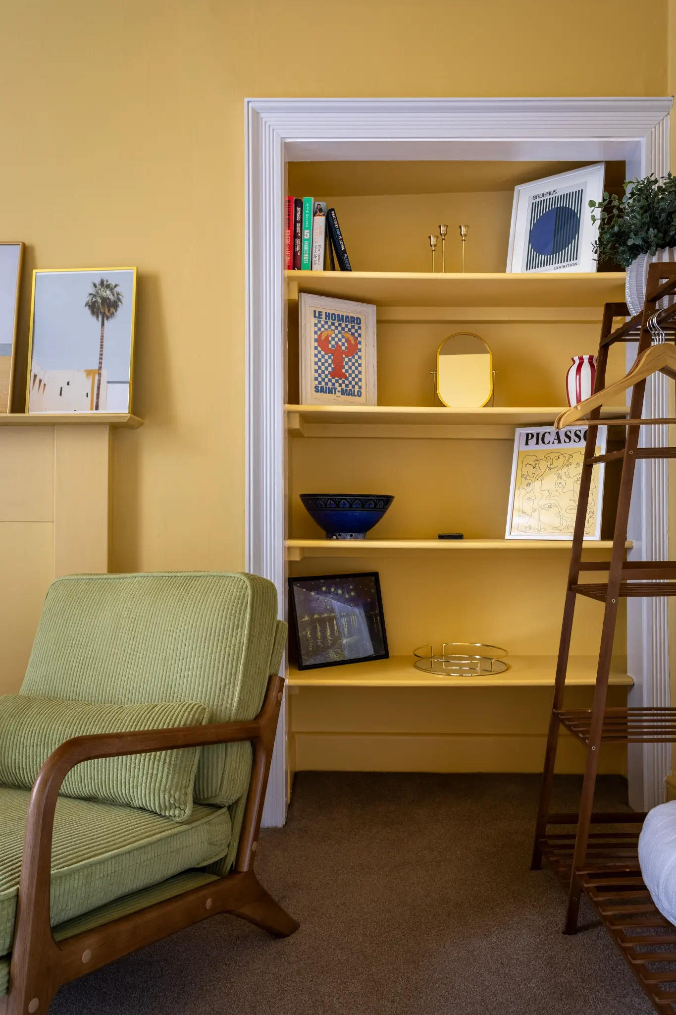 Detail of the amber bedroom bookshelf in a Weymouth esplanade Airbnb with art prints, a green cord armchair, and a wooden clothes rail