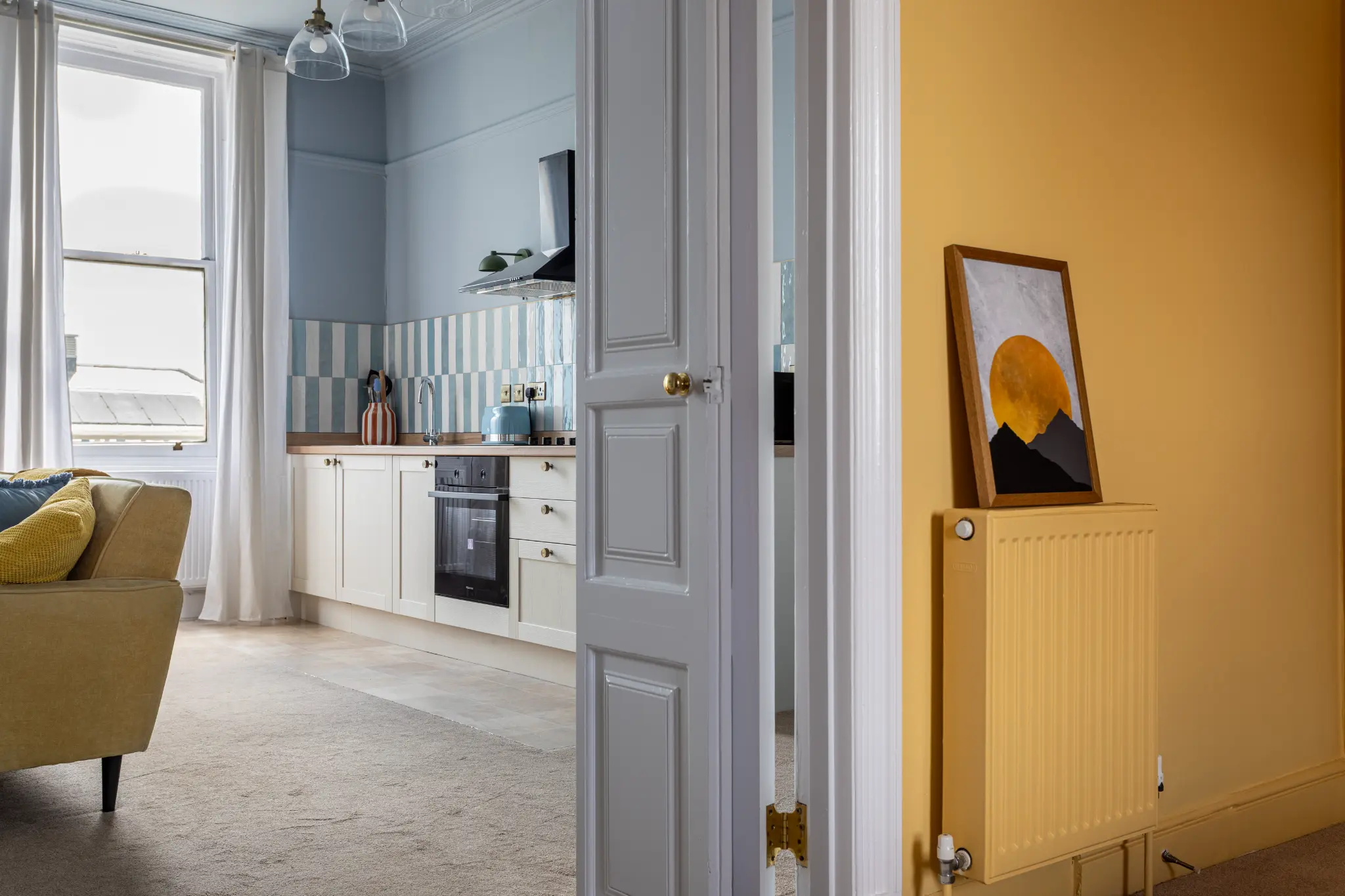 View from the bedroom into the blue living room and kitchen of a Weymouth esplanade Airbnb, showing the transition between the two colour worlds