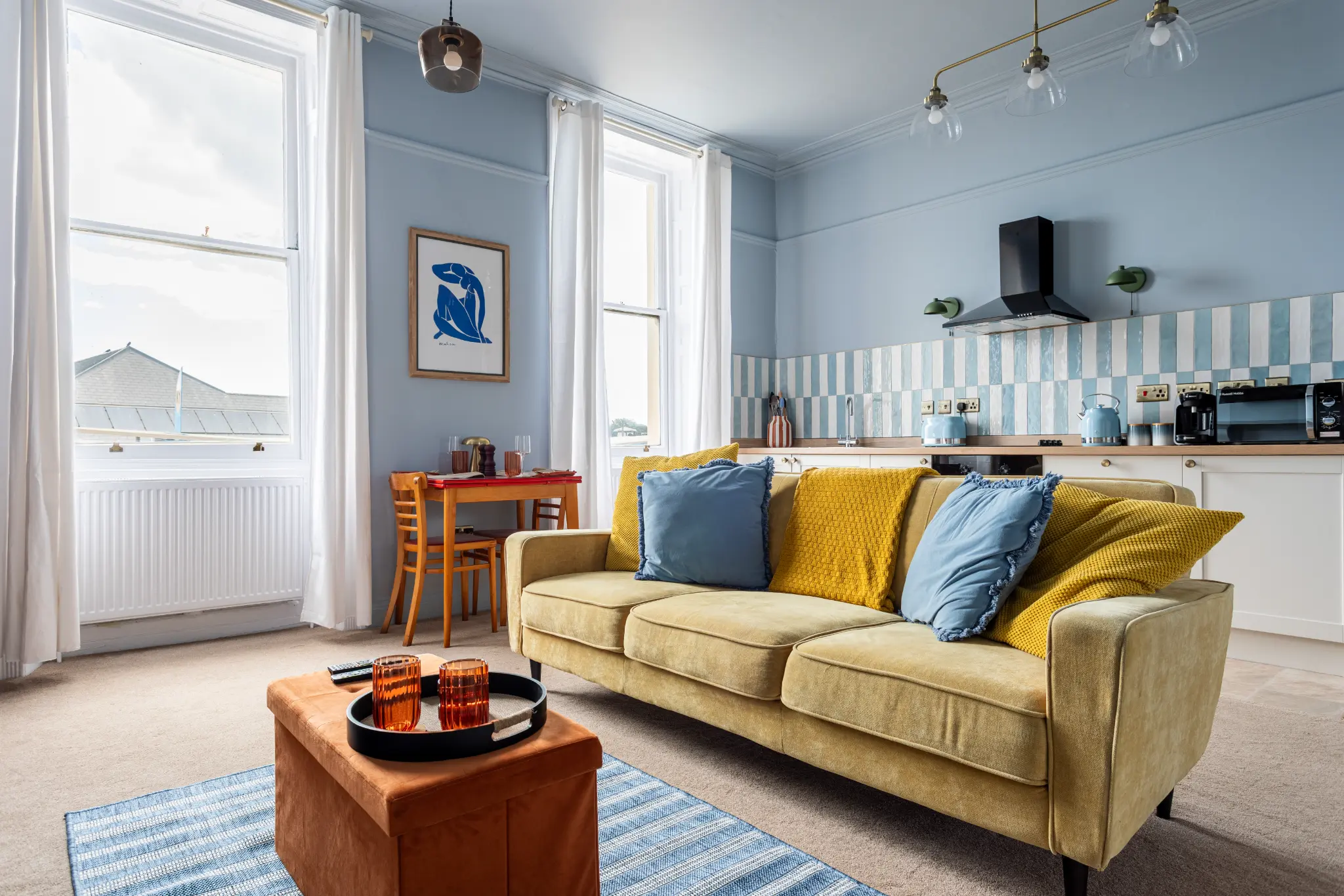 Open-plan living room and kitchen of a Weymouth esplanade Airbnb from the sofa end, showing the chequerboard splashback and esplanade rooftops through the sash windows