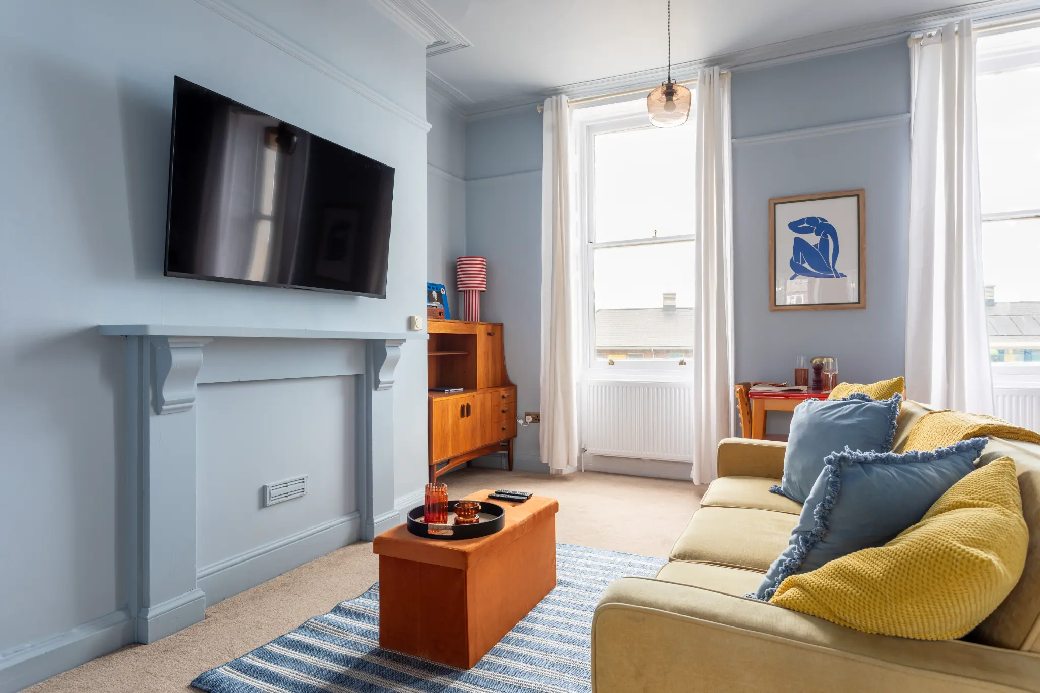 Bohemian Airbnb living room on the Weymouth esplanade showing the mustard sofa, blue cushions, and striped kitchen beyond
